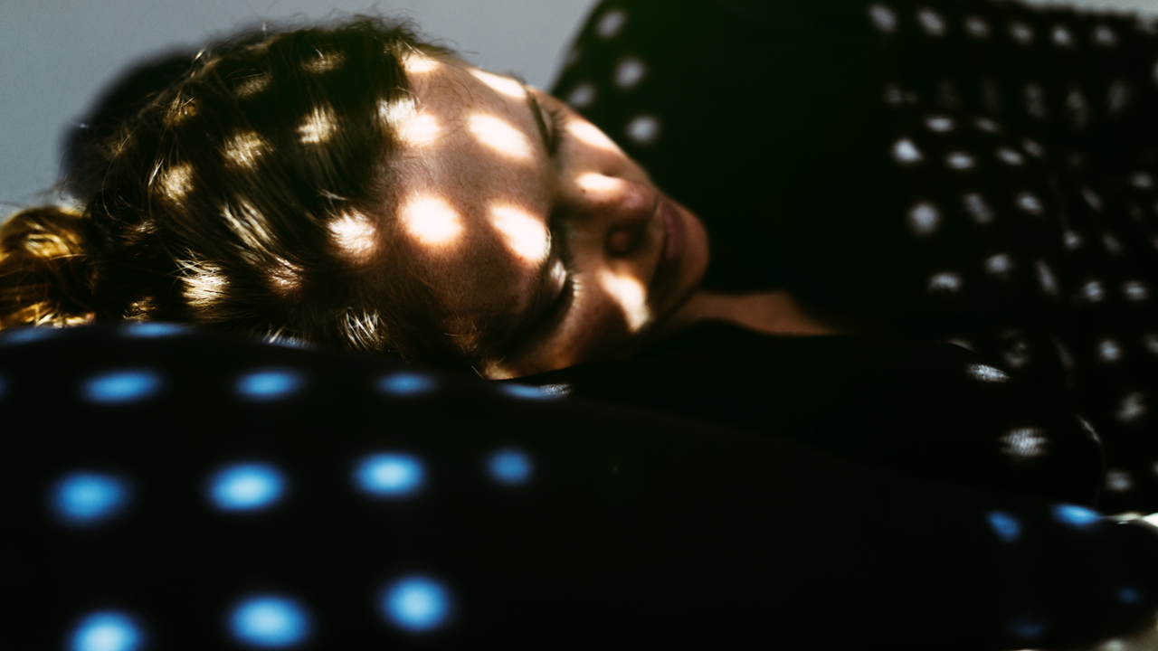 Person asleep with various spots of light casted on their face through gaps in the window blinds.