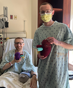 John Nicholas with his kidney donor, Pat Wise, wearing medical gowns in a hospital room and holding toy crocheted kidneys. 