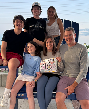 Katie Polkow, Living Well participant and cancer survivor, sits in the middle of her family holding a handmade sign that reads Treatment #16.