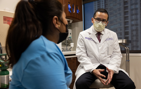 A male Hispanic physician with a white lab coat sitting in an office and talking with a young Hispanic woman who is wearing a light blue shirt.