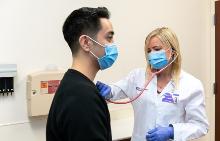 A blonde hair female doctor listening to the heart of a young male patient who is wearing a blue surgical mask.