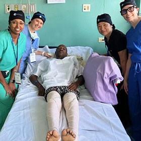 Four female members of a surgical team standing next to a patient who is lying on a hospital bed after a knee replacement.