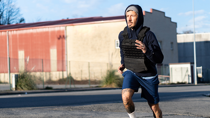 A person wearing a black weighted vest while running outside in shorts.