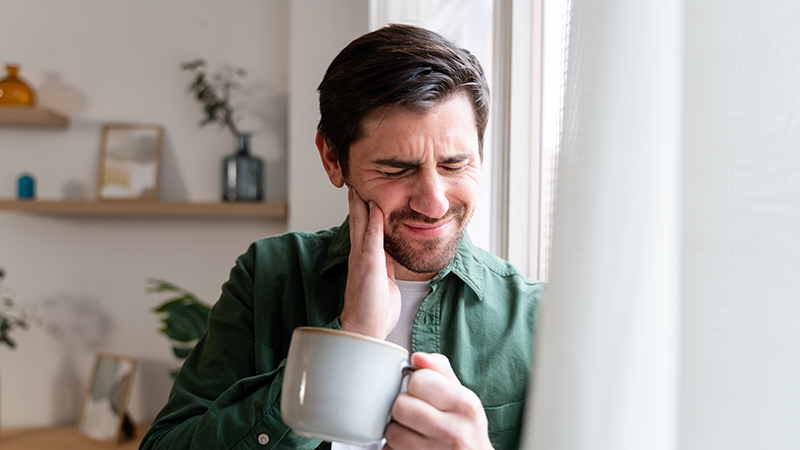 A person holding a cup of coffee and feeling their cheek with their hand.