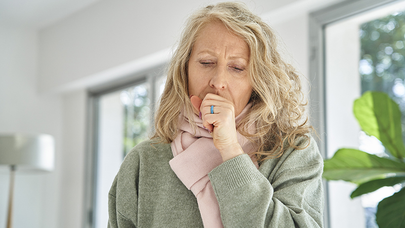 An older adult wearing a green sweater and a pink scarf putting hand on mouth while coughing.