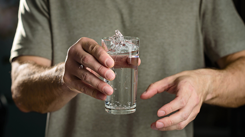 Person pictured from the shoulders down holding a glass of water that looks like it is about to spill.