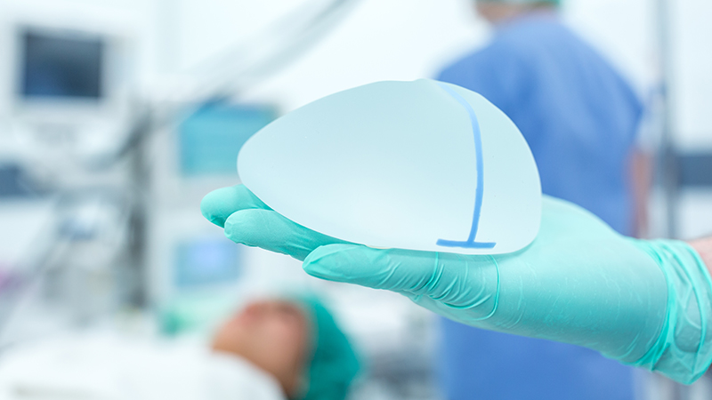 Close up of a hand wearing a blue latex glove holding a breast implant in a surgery room.