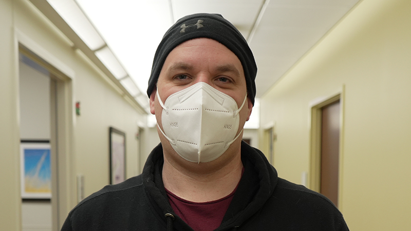 A person in a surgical mask wearing a black hoodie stands in a hospital hallway. 