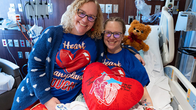 Abbey is on the left side and Meredith is on the right in her hospital bed, and both are smiling wearing “heart transplant tough” shirts. 